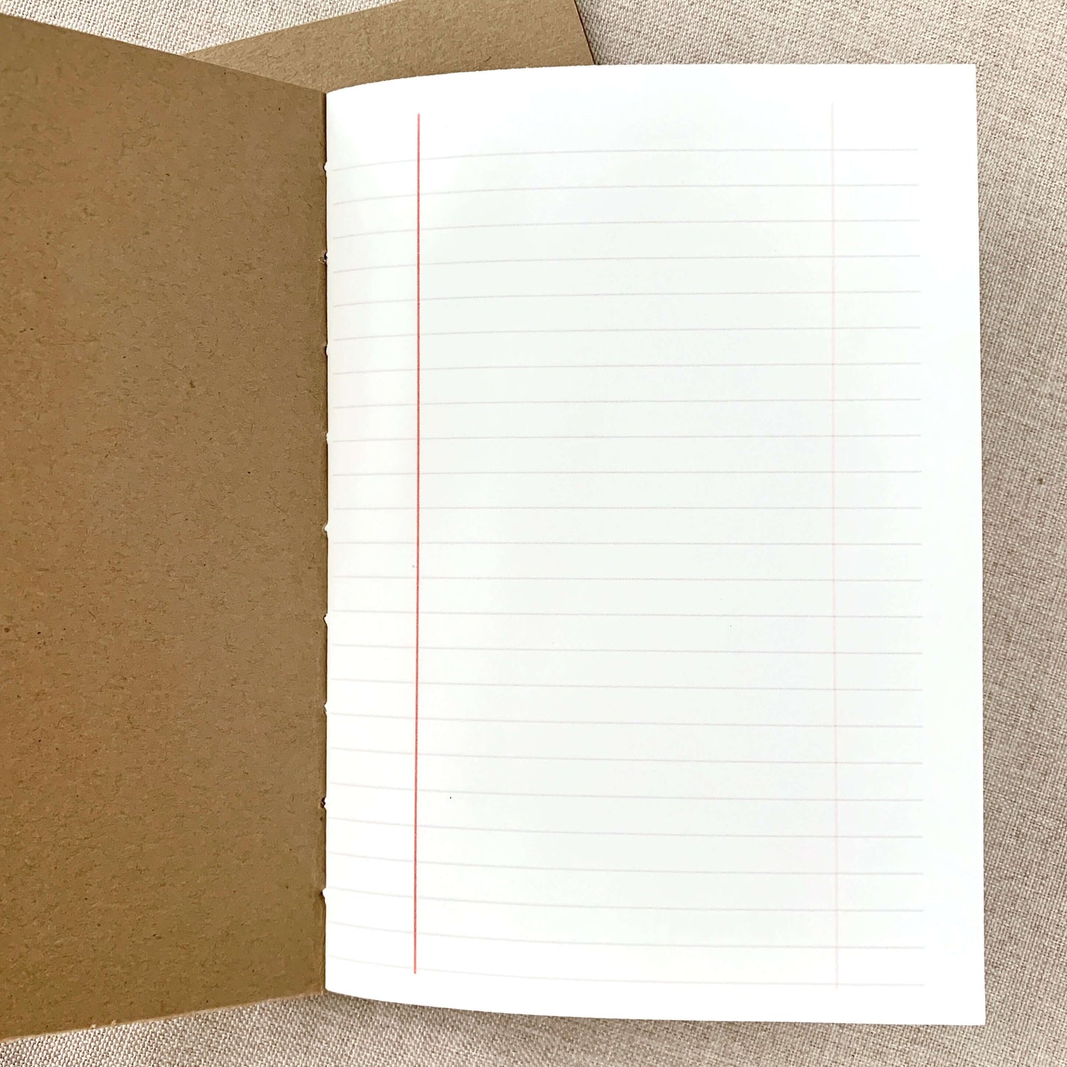 Open notebook showing lined white paper with a red margin line inside a hand-stitched kraft cover, labeled A6 Composition Lined Travelers Notebook Insert - Fountain Pen Friendly, Hand-Stitched Kraft Cover